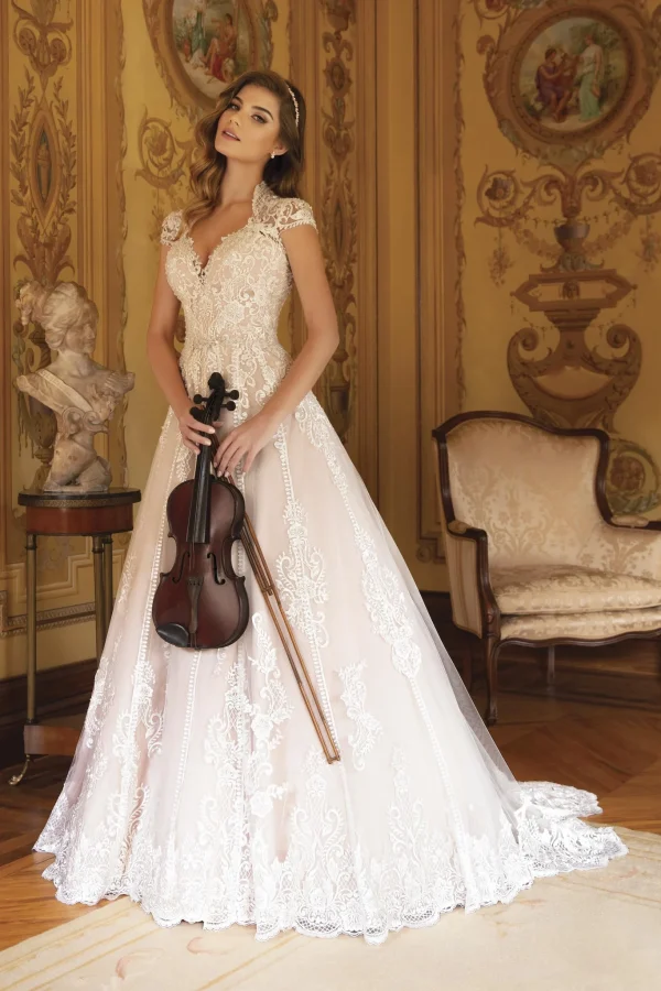 modella indossa un abito da sposa color cipria con fitti ricami in pizzo bianco, scollo a V e spalline decorate, posando in una sala d'epoca con un violino