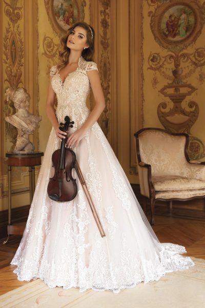 modella indossa un abito da sposa color cipria con fitti ricami in pizzo bianco, scollo a V e spalline decorate, posando in una sala d'epoca con un violino